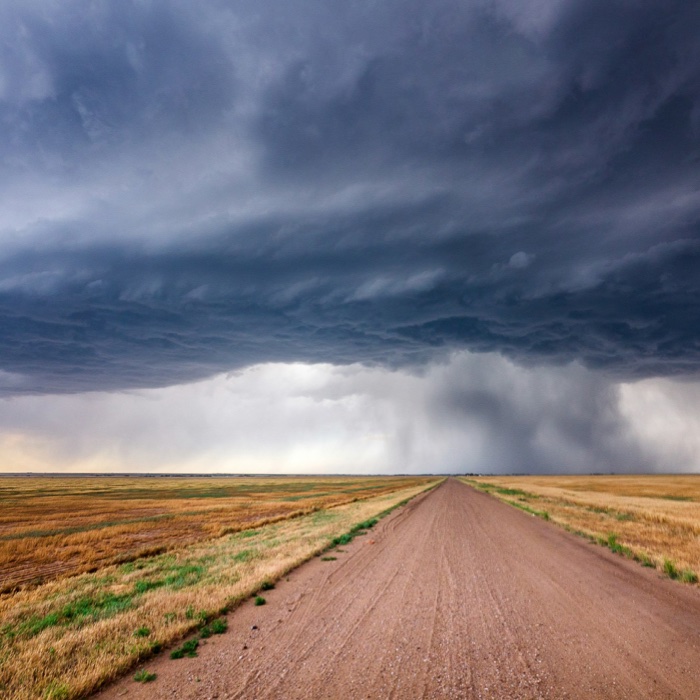 A dirt road running to the horizon under a layered storm sky over open prairie.